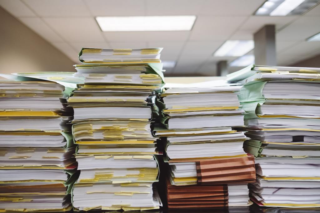 View of four high stacks of documents and files on a table in an office environment. The viewer is on eye level with the stacks so those piles of documents feel overwhelming