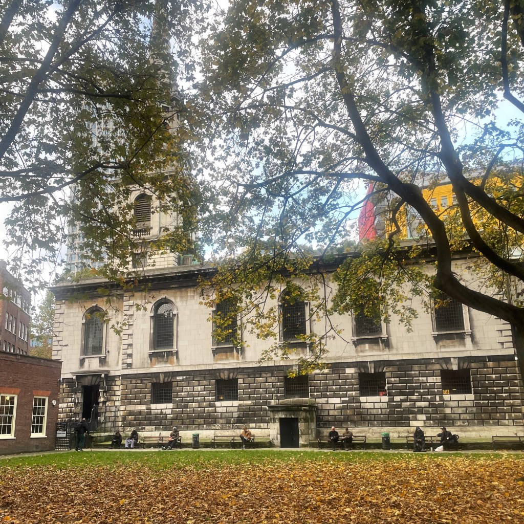 St Giles-in-the-Fields viewed from the churchyard. The gras in the foreground is covered with brown leaves, the church spire and modern buildings behind the church are partly abscured by trees.