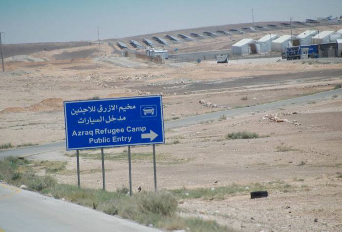 In a desert landscape, a blue sign on the side of the road that reads in both Arabic and English: Azraq Refugee Camp Public Entry.