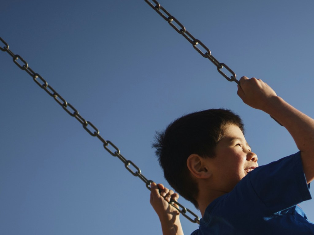 A young boy on a swing photographed from below against a blue sky. We only see his head and hands holding on to the swings chains. We see him from behind, his head turned to the right, his face in profile. he clearly enjoys the swing.